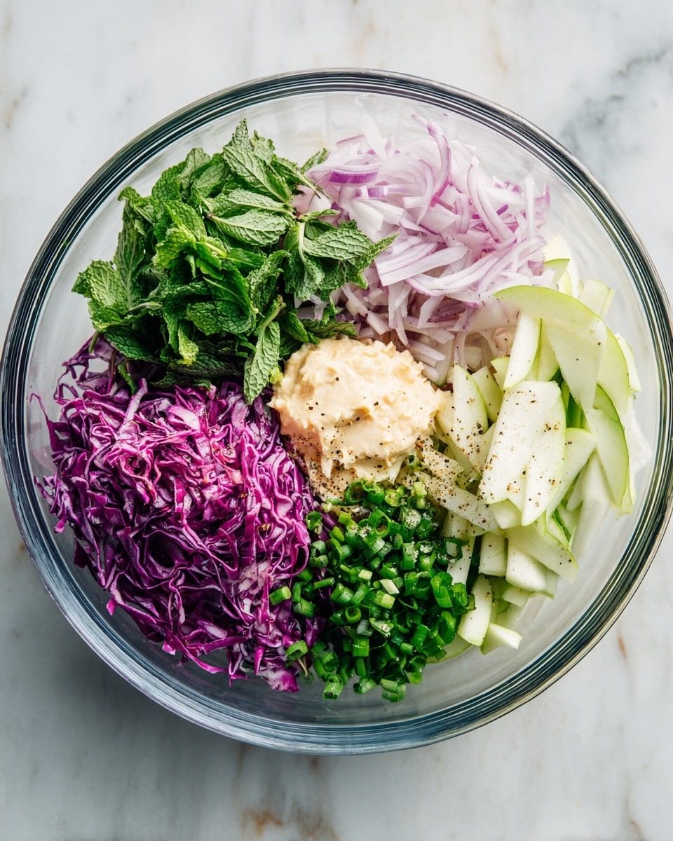 A clear glass bowl on a white marbled surface holds several colorful layers of fresh ingredients, each section separated from the others. On the left, there is a thick layer of bright purple shredded cabbage with a rough texture. Above and next to it, nicely shredded green mint leaves add a fresh touch. At the upper center, very thin slices of light purple and white onions form a soft, slightly translucent mound. On the right side, pale green thin apple strips with a smooth texture are stacked in a small pile. In the middle, a creamy beige dollop of sauce sits on top of a mix of chopped dark green herbs and black pepper, with small dots of white salt and fresh green chopped scallions surrounding it. The overall look is fresh, colorful, and organized. Photo taken with an iphone --ar 4:5 --v 7