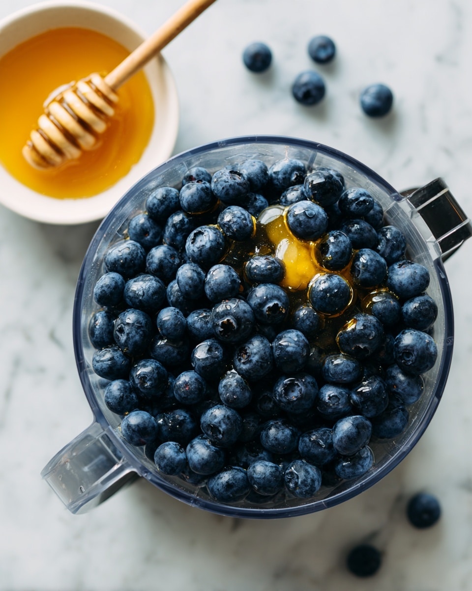 A clear food processor bowl filled with many fresh, dark blue blueberries sitting closely together with a shiny, small layer of golden honey drizzled over them. A few blueberries are scattered on the white marbled surface around the processor. At the top left corner, there is a small white bowl filled with thick golden honey and a honey dipper resting inside it, partly covered with honey. The scene shows a top-down view, with soft natural light highlighting the textures and colors. Photo taken with an iphone --ar 4:5 --v 7