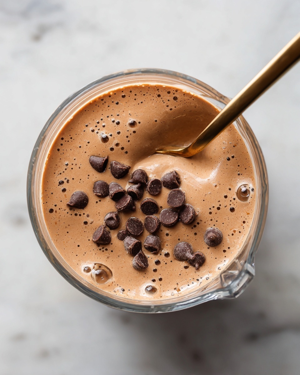 The image shows a close-up of a creamy chocolate smoothie inside a clear blender jar. The smoothie is light brown with a smooth texture and small bubbles on the surface. Scattered on top are dark chocolate chips that add texture and contrast in color. A gold metal spoon is scooping some of the smoothie from the jar, resting on the right side of the image. The jar is placed on a white marbled surface. photo taken with an iphone --ar 4:5 --v 7