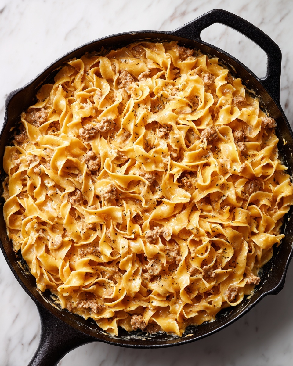 A close-up view of a black cast iron pan filled with creamy pasta noodles mixed with a brownish-orange sauce. The dish has wide, flat noodles that are twisted and layered over each other, coated evenly in the thick sauce. Small pieces of light brown protein are scattered throughout the pasta, adding texture contrast. The pan is placed on a white marbled surface, creating a clean and bright background. photo taken with an iphone --ar 4:5 --v 7