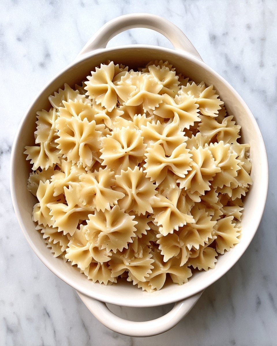 A white round ceramic dish with two small handles is filled with cooked bow-tie pasta. The pasta is light beige in color with a smooth texture and sits evenly spread out inside the dish. The background is a white marbled surface, contrasting with the dish and pasta. photo taken with an iphone --ar 4:5 --v 7