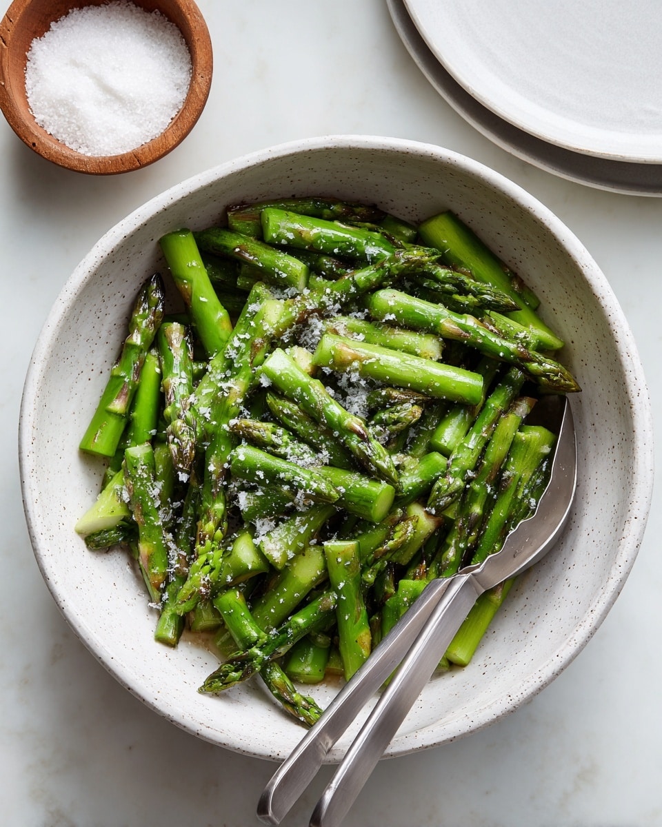 A white speckled bowl holds bright green asparagus pieces, all cut into short sections with some tips showing a slight purple tint. The asparagus is sprinkled with coarse salt, giving a grainy texture on top. A pair of metal tongs with gray silicone tips lies diagonally across the bowl, partially resting on the asparagus. In the top left corner, a small white bowl filled with coarse salt is visible on a white marbled surface. photo taken with an iphone --ar 4:5 --v 7