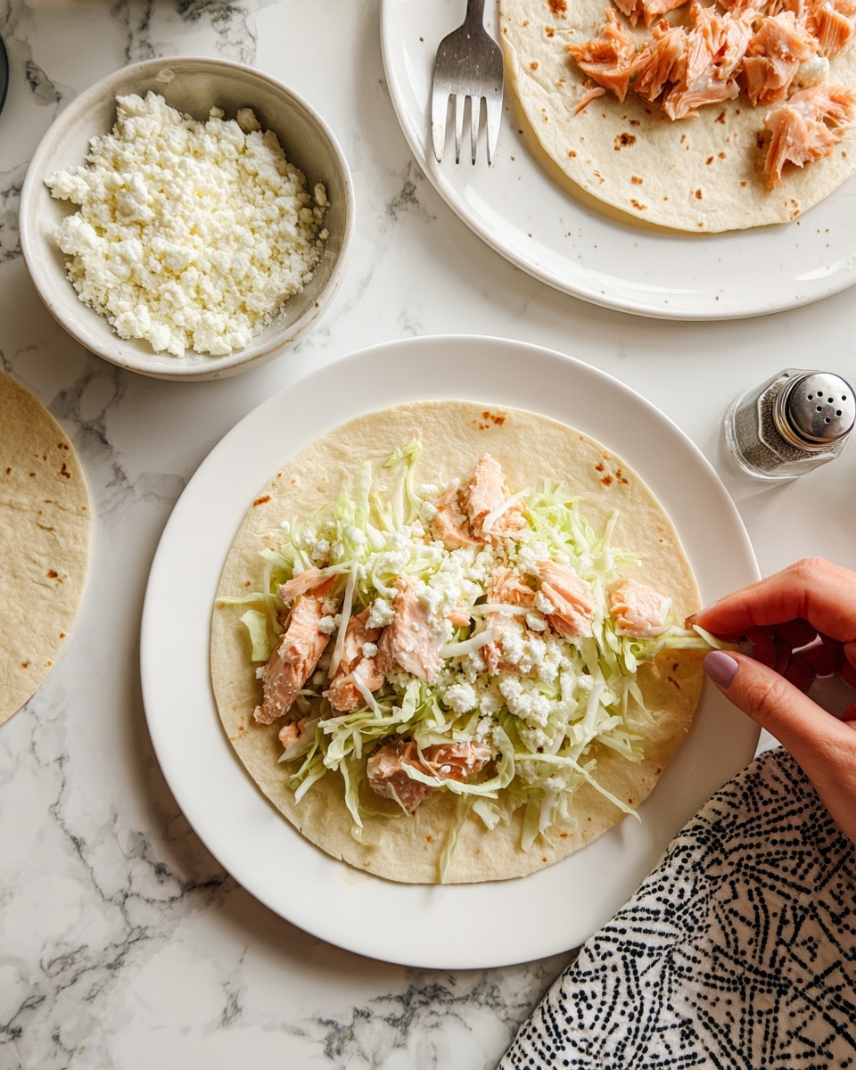 The image shows a white plate with a soft, pale yellow tortilla as the base. On the tortilla, there are layers of pink cooked fish chunks scattered evenly, topped with white crumbly cheese, and a layer of light green shredded cabbage being placed by a woman's hand. To the side, there is another white plate with a tortilla topped with similar ingredients. Above, a white plate with flaky pink fish pieces and a silver fork is visible. On the white marbled surface, a small bowl filled with white crumbly cheese and a pepper shaker are also placed. A patterned cloth with black line designs is partly visible under the plates. photo taken with an iphone --ar 4:5 --v 7