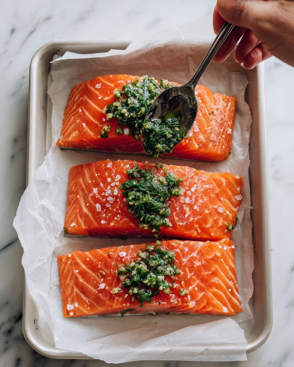 Salmon and Asparagus Green Goddess Bowl Recipe 4 The image shows three thick, bright orange-red pieces of raw salmon with a flaky texture, laid out on white parchment paper inside a white tray. Each piece is sprinkled with coarse salt, making small white grains visible on the surface. Over the middle piece, a silver spoon held by a person's woman's hand is placing a green herb sauce with visible dill and minced garlic, adding a fresh and textured topping. The background features a white marbled surface, giving a clean and simple look. photo taken with an iphone --ar 4:5 --v 7