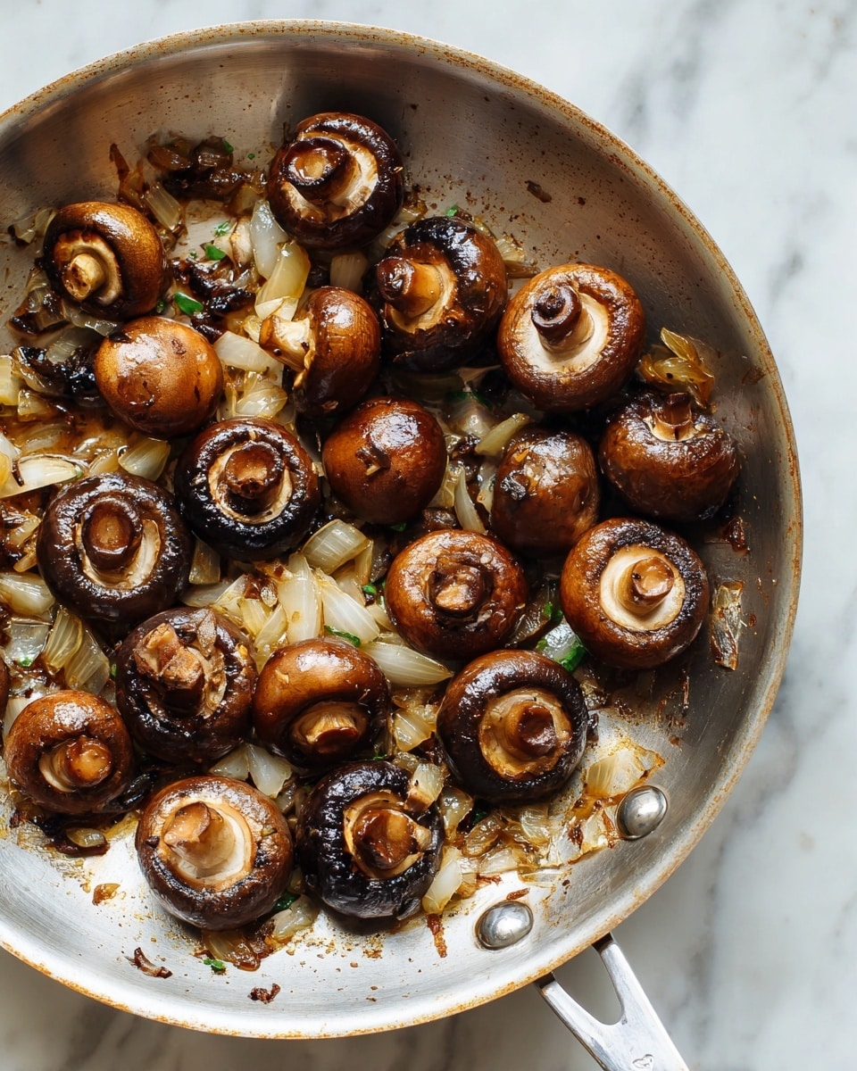 The image shows a frying pan filled with cooked whole brown mushrooms, some turned upside down to show their dark gills, mixed with small pieces of golden-brown sautéed onions. The mushrooms have a shiny, slightly caramelized surface with a light brown color, and the pan has a few browned bits stuck to the bottom, showing a rich cooking process. The pan is silver metal with two rivets near the bottom edge and a handle partly visible on the left. The background is a white marbled texture. photo taken with an iphone --ar 4:5 --v 7