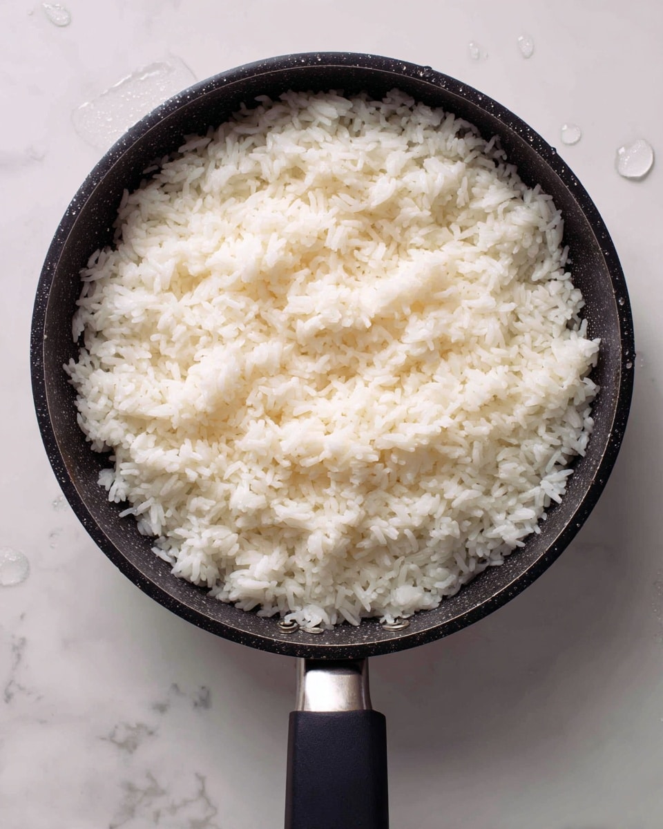 A close-up top view of a black strainer filled with cooked white rice, showing small, soft, and slightly sticky rice grains that have a light sheen. The strainer rests on a white marbled surface, with droplets of water on the black handle and edges of the strainer. The rice layer is thick and evenly spread, filling the entire round strainer. photo taken with an iphone --ar 4:5 --v 7