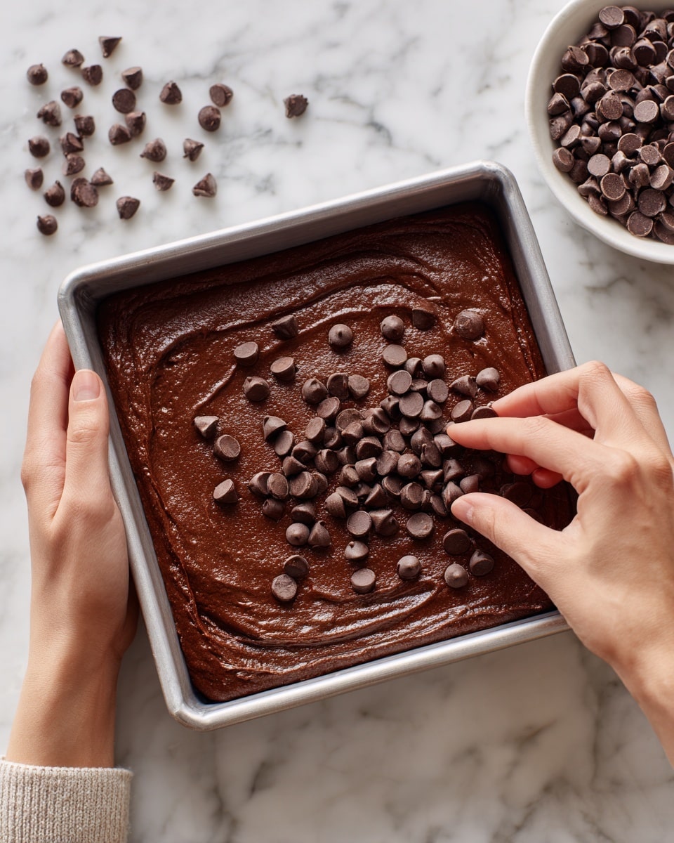 A square metal baking pan filled with a smooth, thick, dark brown chocolate batter spread evenly in one layer. Small dark chocolate chips are sprinkled on top in a scattered pattern. A woman's hand is gently placing more chocolate chips on the batter from the right side, while another woman's hand holds the pan from the left side. In the top right corner, a white round dish contains more chocolate chips, and the scene is set on a white marbled surface. photo taken with an iphone --ar 4:5 --v 7