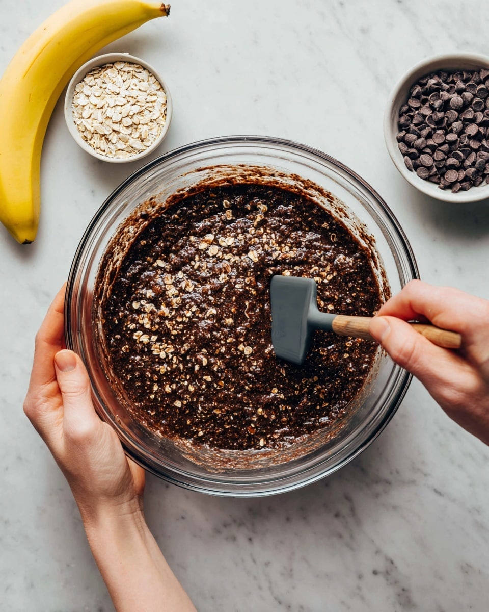 A clear glass bowl is filled with a thick, dark brown mixture loaded with visible light brown oats spread evenly throughout. The mixture has a glossy wet texture with a slightly chunky look from the oats. A woman's hand holds the bowl steady on the left side, while another woman's hand stirs the mixture with a dark gray spatula on the right side. Above the bowl, on a white marbled surface, sits a small white bowl filled with dark brown chocolate chips and a ripe yellow banana nearby. photo taken with an iphone --ar 4:5 --v 7