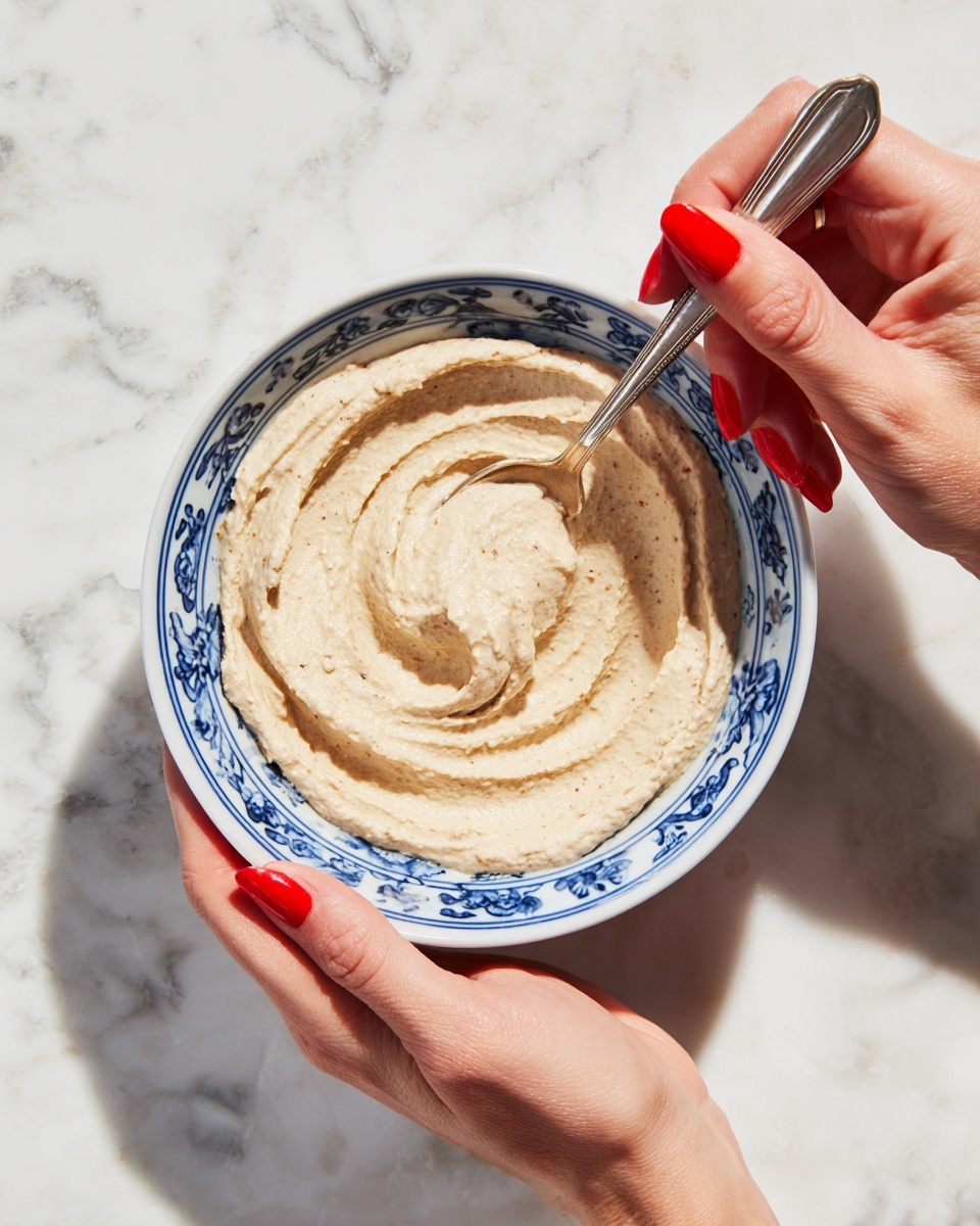 A white bowl with blue patterns holds a smooth, creamy beige dip spread in a round shape. A woman's hand with bright red polished nails holds the bowl on the left side, while the other woman's hand uses a silver spoon to swirl the dip in the center. The dip has a thick, soft texture with small specks inside. The background is a white marbled texture photo taken with an iphone --ar 4:5 --v 7