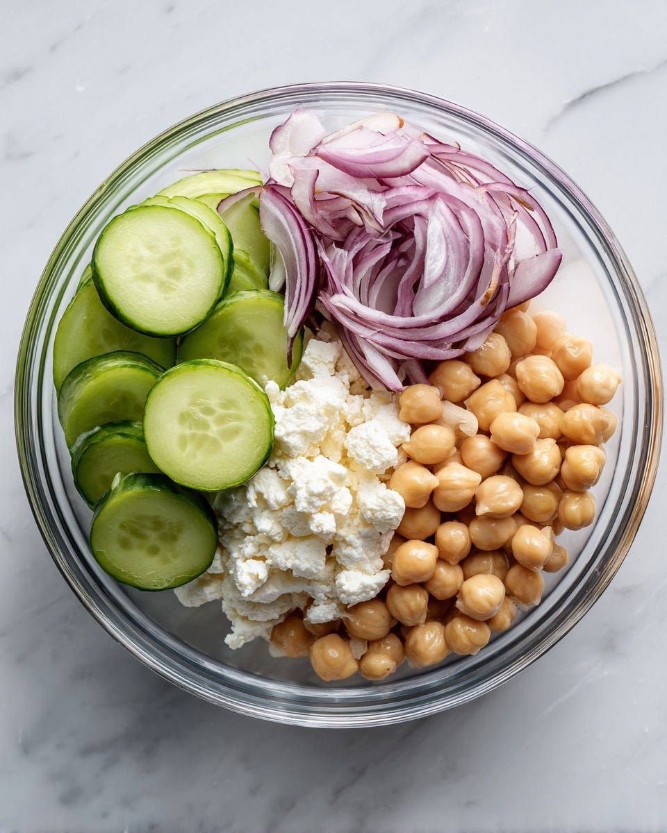 A clear glass bowl sits on a white marbled surface, holding four distinct layers inside. On the left side, there are bright green cucumber slices arranged in a neat pile, showing their smooth, round shapes. Next to them is a small bunch of thin, pale purple sliced shallots with a slightly curled texture. Below the shallots, there is a mound of soft, crumbly white cheese that covers part of the chickpeas beneath it. The bottom right section is filled with smooth, light brown chickpeas, rounded and slightly shiny, forming the base of the bowl. photo taken with an iphone --ar 4:5 --v 7
