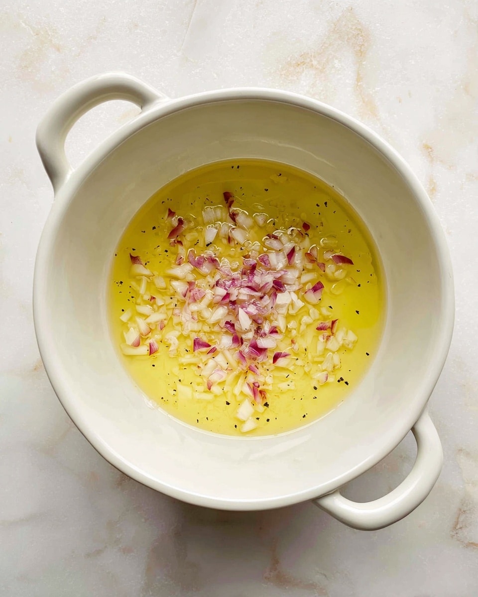 A white pot with two handles viewed from above shows a shallow layer of light yellow oil with small pieces of finely chopped red and white shallots scattered in the center. The background is a white marbled texture. Photo taken with an iphone --ar 4:5 --v 7