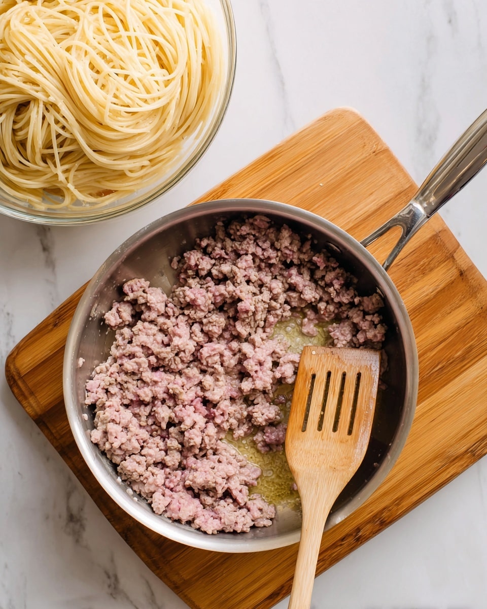 A round silver pan sits on a wooden board with cooked ground meat that is partly brown and partly pink, showing it is still cooking, mixed with a light layer of oil at the bottom. A light-colored wooden spatula rests inside the pan on the right side. To the left of the pan, there is a transparent bowl full of plain, cooked spaghetti noodles with a pale yellow color. The whole scene is set on a white marbled surface. photo taken with an iphone --ar 4:5 --v 7