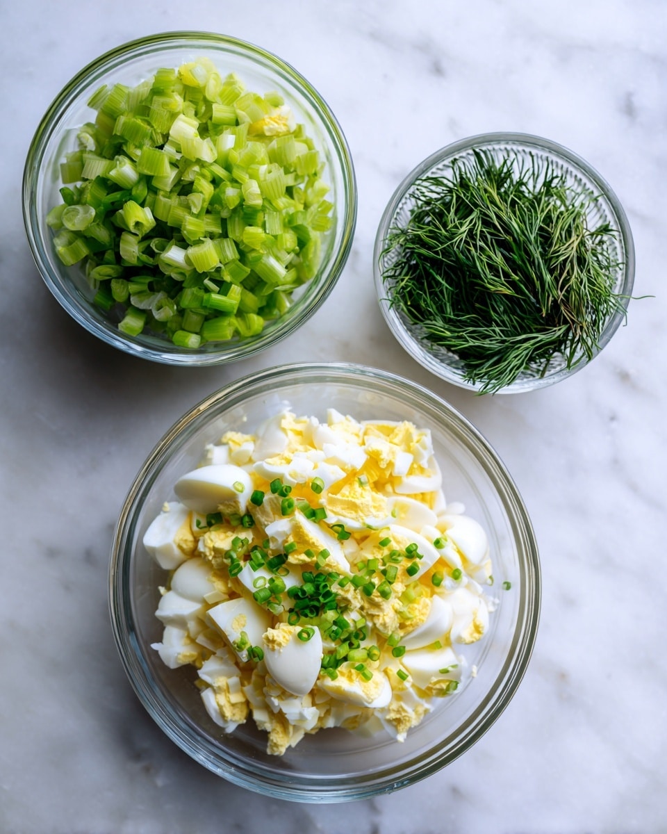 The image shows three clear glass bowls on a white marbled surface. The largest bowl at the bottom contains chopped hard-boiled eggs with white and yellow pieces, along with small green chopped chives on top. To the top left is a medium bowl filled with finely chopped light green celery. To the top right is a small bowl holding thin, dark green dill sprigs. The arrangement is simple and clean, with a fresh and natural look. photo taken with an iphone --ar 4:5 --v 7