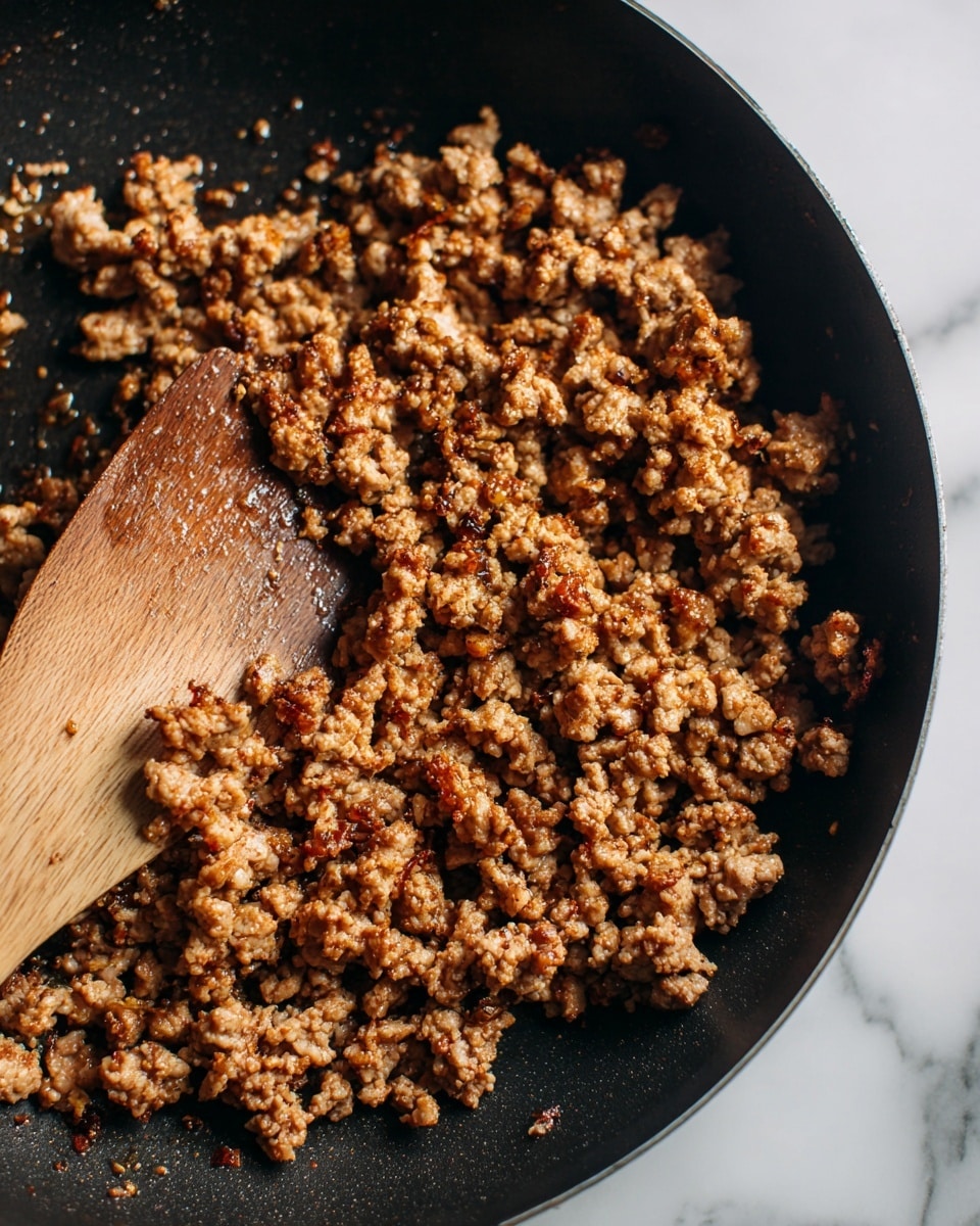 This image shows a close-up of cooked ground meat in a black frying pan. The meat is broken into small, uneven pieces with a mix of light brown and darker crispy brown colors. There are small bits that look slightly crunchy, and the texture is crumbly but moist. A wooden spatula is resting in the pan with some bits of meat stuck to it. The background is a white marbled surface. photo taken with an iphone --ar 4:5 --v 7