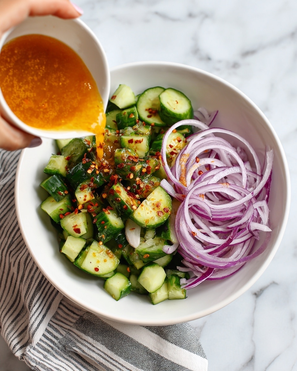 A white bowl filled with two main layers: the bottom layer is light purple thin onion rings, and the top layer is small green cucumber cubes with dark green skin. A woman's hand is pouring a bright orange dressing with red pepper flakes over the cucumbers and onions. The bowl is on a white marbled surface with a striped cloth nearby. Photo taken with an iphone --ar 4:5 --v 7