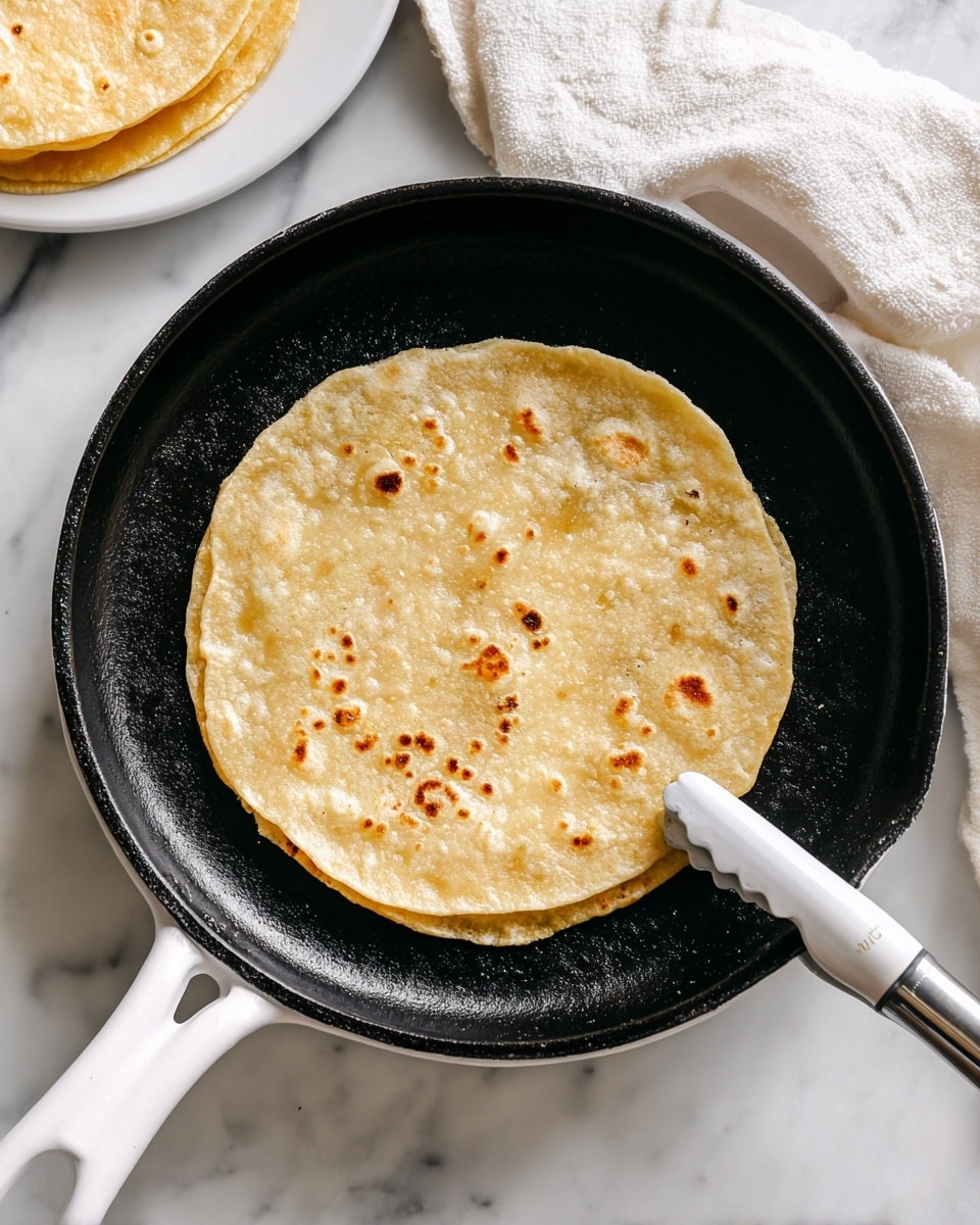 A single round, light golden tortilla with small bubbles and an uneven surface is frying in a black cast iron pan with a white handle. The tortilla lies flat in some oil, showing a slight sheen, while a white kitchen tong touches it gently from the right side. In the top left corner, there is a glimpse of a white plate holding another tortilla on a white napkin. The scene is set on a white marbled surface. Photo taken with an iphone --ar 4:5 --v 7