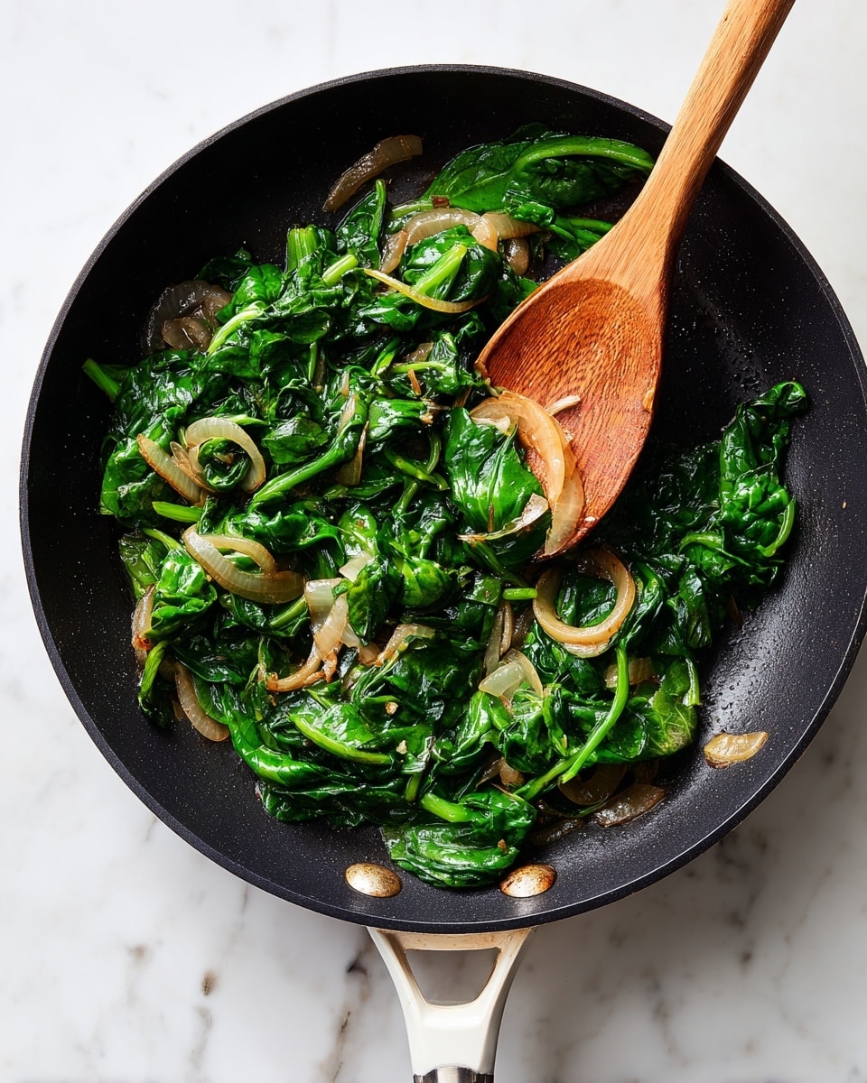 A black frying pan with a white handle sits on a white marbled surface, filled with sautéed spinach and light brown, soft-looking onion rings spread evenly. The spinach leaves are dark green with a slightly wilted texture, mixed with translucent, cooked onions. A wooden spoon stirs the vegetables gently, its natural wood grain clear and smooth, resting inside the pan. The overall look is fresh and simple with a natural color contrast between the green spinach and pale onions. photo taken with an iphone --ar 4:5 --v 7