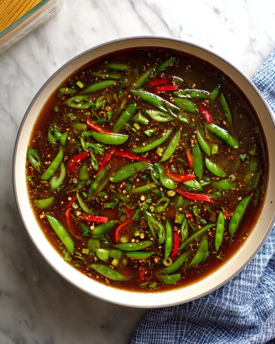 The image shows a white pan filled with a dark brown broth full of bright green snap peas and thin red strips of bell peppers floating evenly throughout the liquid. The broth has visible spices speckled across the surface, giving it a textured look. The pan is placed on a white marbled surface, with a small part of a white container holding uncooked spaghetti visible at the top left. To the right, a blue and white checkered cloth adds a soft touch to the scene. Photo taken with an iphone --ar 4:5 --v 7