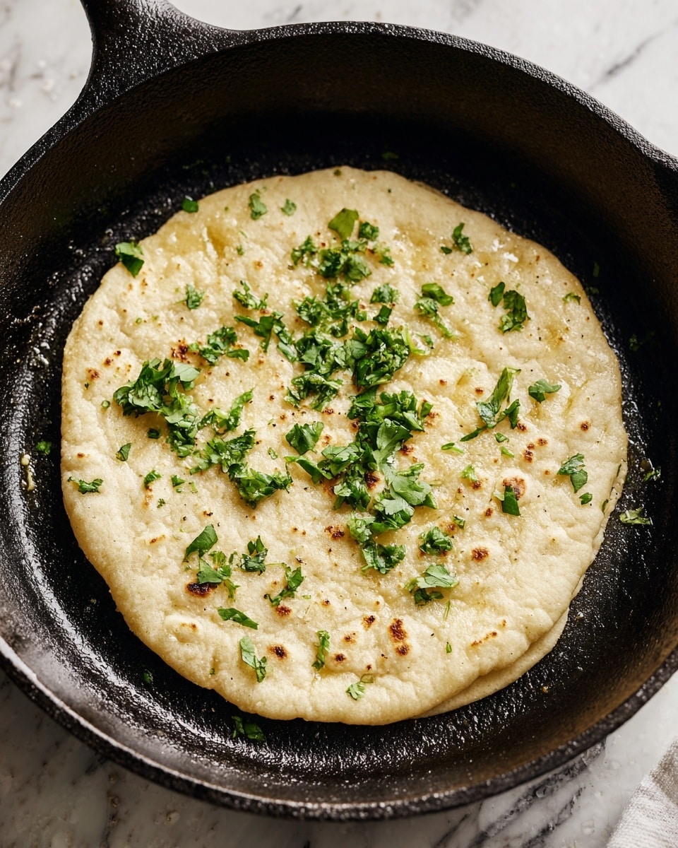 A small, round flatbread is cooking in a black cast iron skillet. The flatbread has a light beige color with a smooth texture and some small air bubbles forming on the surface. On top, there are scattered pieces of green herbs, likely cilantro or parsley, adding a fresh touch. The skillet is centered on a stovetop burner with a white marbled surface visible around it. photo taken with an iphone --ar 4:5 --v 7
