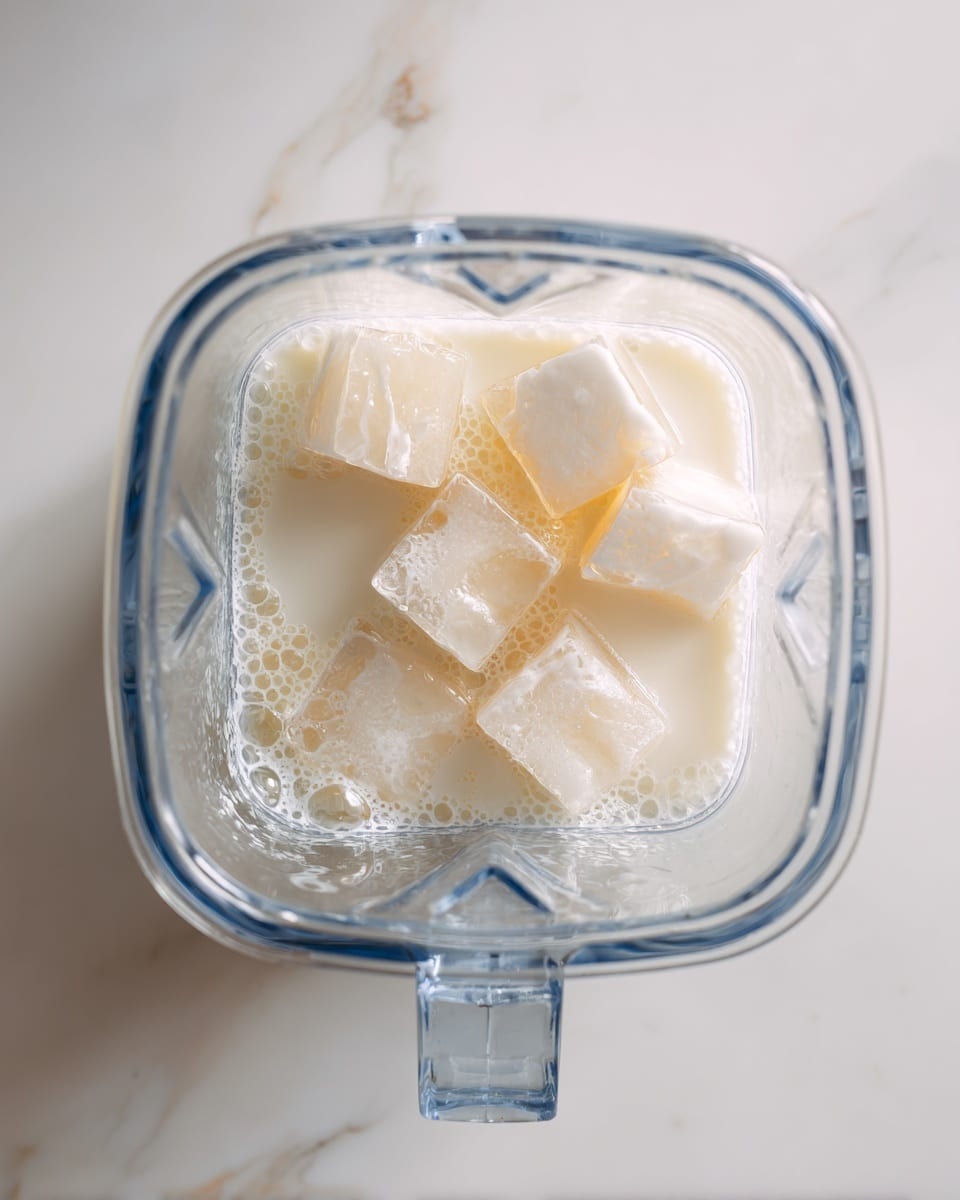 The image shows a clear blender container on a white marbled surface. Inside the container, there is a single layer of white milk at the bottom, with several light yellowish ice cubes floating on the milk. Small bubbles are scattered mainly on one side of the milk’s surface. The container is square-shaped and viewed from the top, with a visible handle on one corner. Photo taken with an iphone --ar 4:5 --v 7