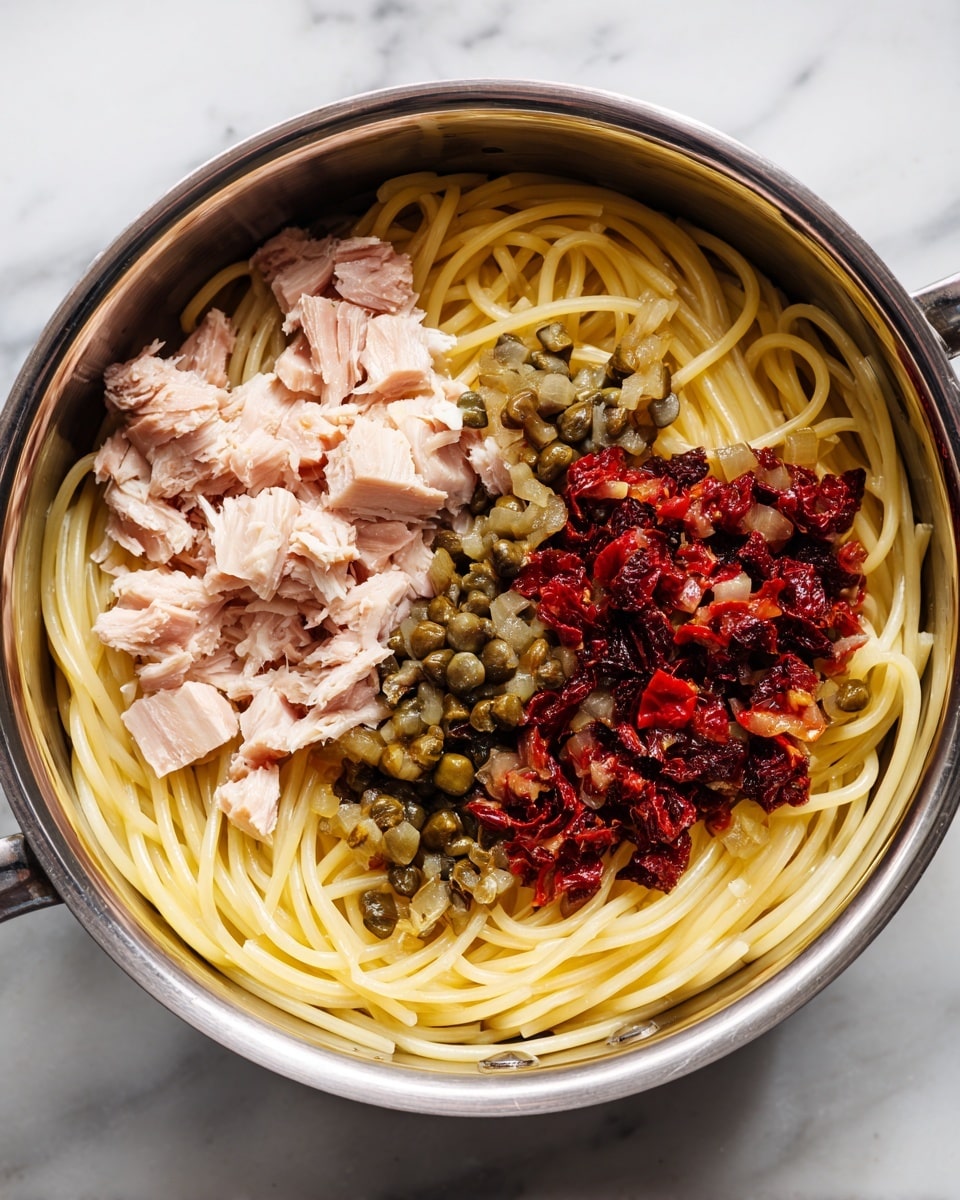 A shiny metal pot contains three layers of ingredients: the bottom layer is long, thin pale yellow spaghetti, loosely spread across the pot; on the left side, on top of the pasta, there are chunks of light pink tuna with a soft texture; on the right side, there is a colorful mix of small green capers and chopped dark red sun-dried tomatoes mixed with small bits of garlic or onion, creating a textured, varied surface. The pot sits on a white marbled surface. photo taken with an iphone --ar 4:5 --v 7