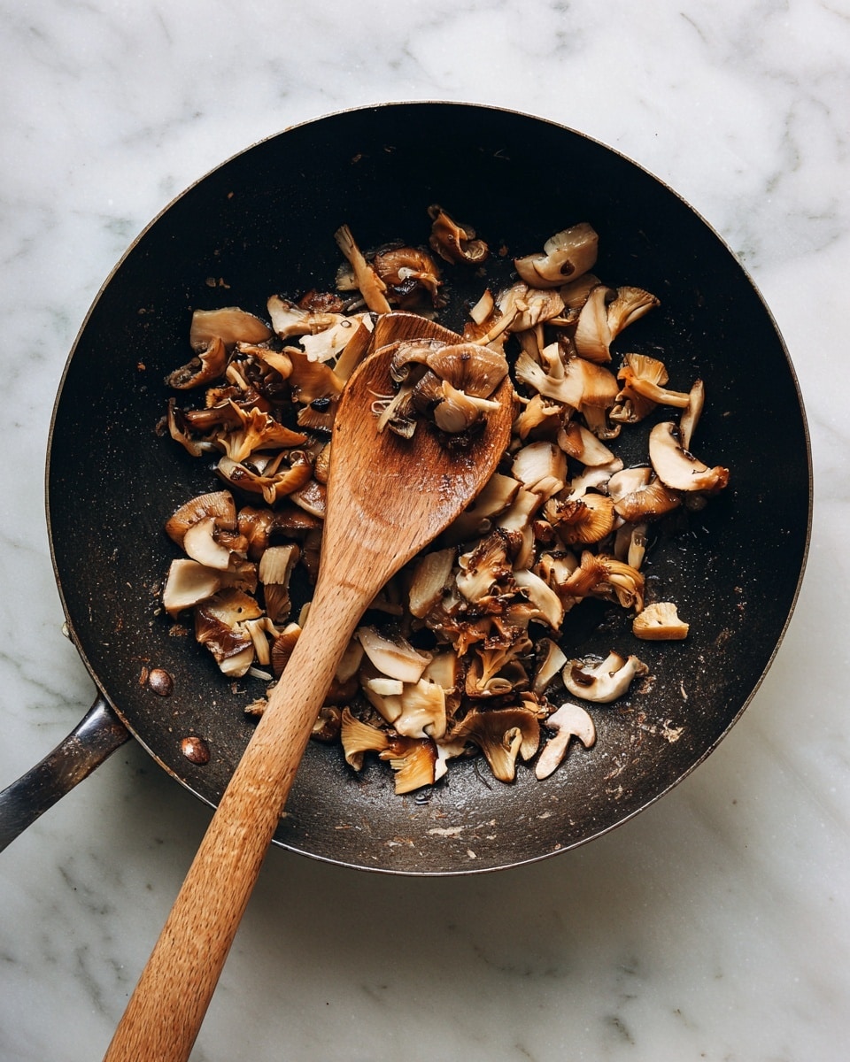 A dark, slightly worn pan filled with small pieces of light brown mushrooms being cooked. The mushrooms are scattered and look soft with some slightly browned edges. A wooden spoon with a long handle rests inside the pan, touching some mushrooms. The pan is placed on a white marbled surface. Photo taken with an iphone --ar 4:5 --v 7