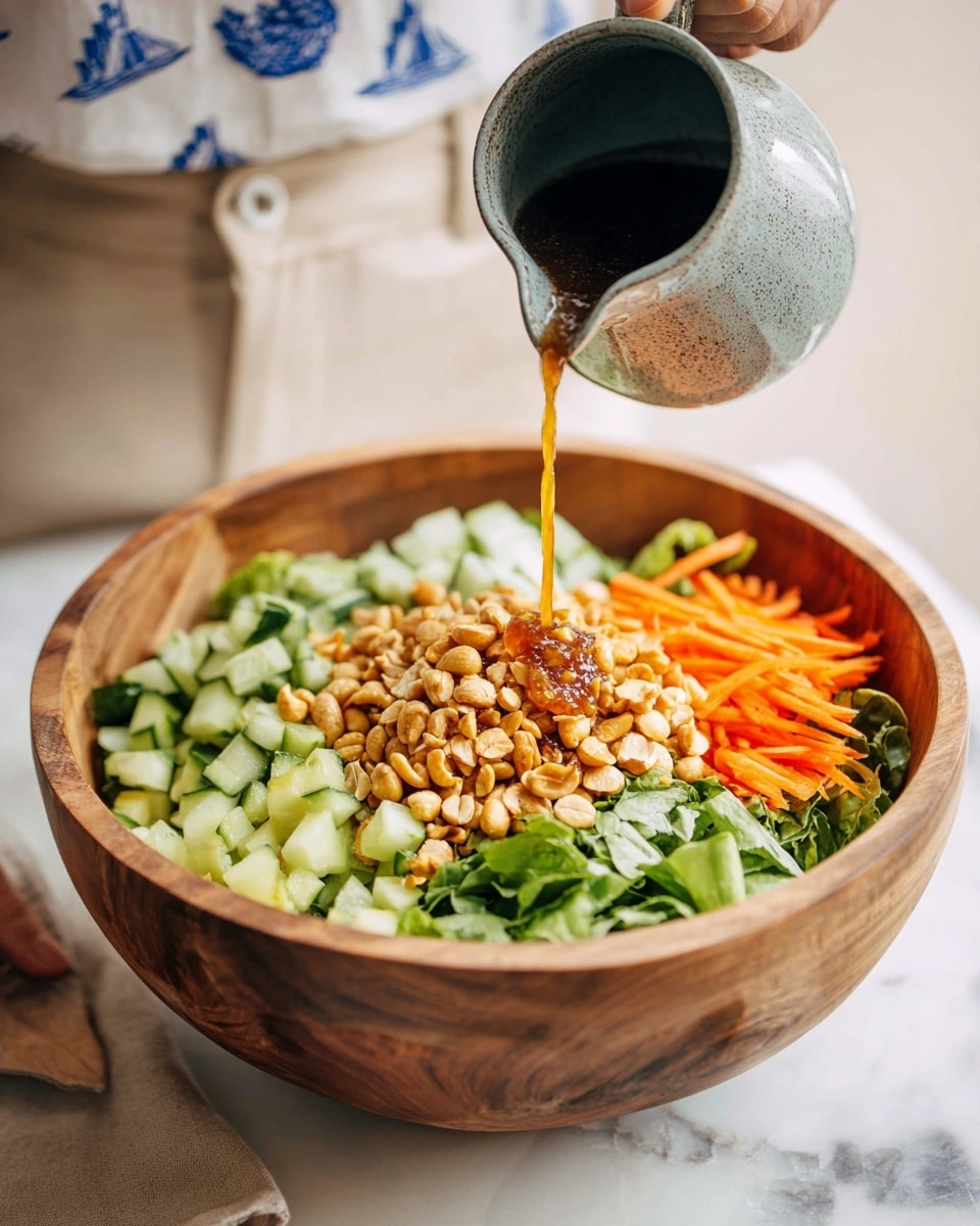 A wooden bowl filled with a layered salad sits on a white marbled surface. The bottom layer appears to be chopped green leafy vegetables, topped with small diced pale green cucumber pieces, thin long orange carrot sticks, and a generous pile of golden-brown roasted nuts in the center. A woman's hand holds a grey ceramic pitcher, pouring a thin stream of brown dressing onto the nuts. The person in the background wears light beige pants and a white shirt with blue floral and nautical patterns. Photo taken with an iphone --ar 4:5 --v 7